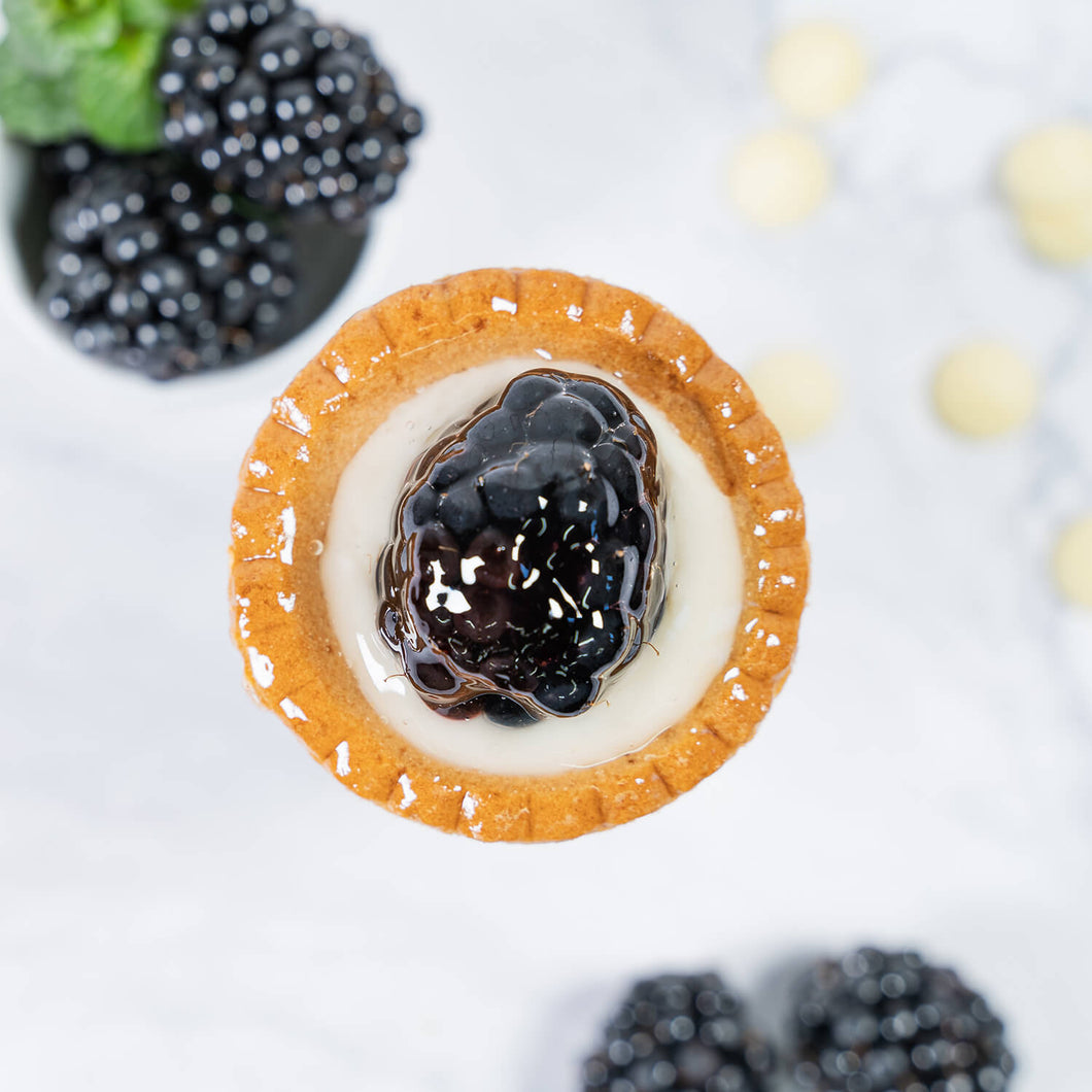 Brombeer Tartelette aus Mürbteig, Creme und mit einer Brombeere belegt, zur Deko frische Brombeeren und weiße Schokoladenstückhen